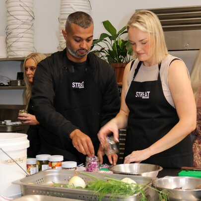 Two people wearing aprons preparing food on a bench in a kitchen setting.