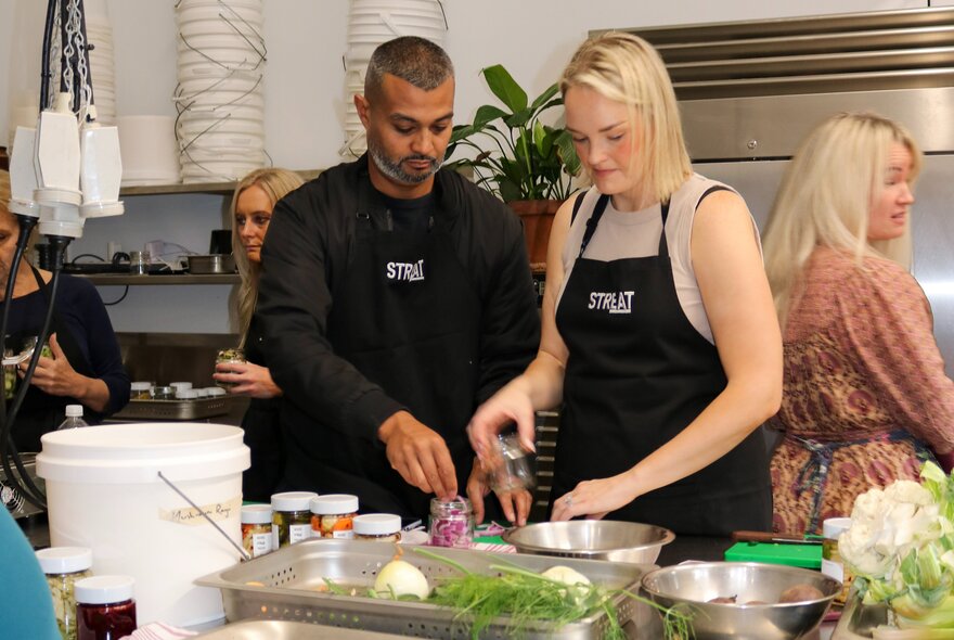 Two people wearing aprons preparing food on a bench in a kitchen setting.
