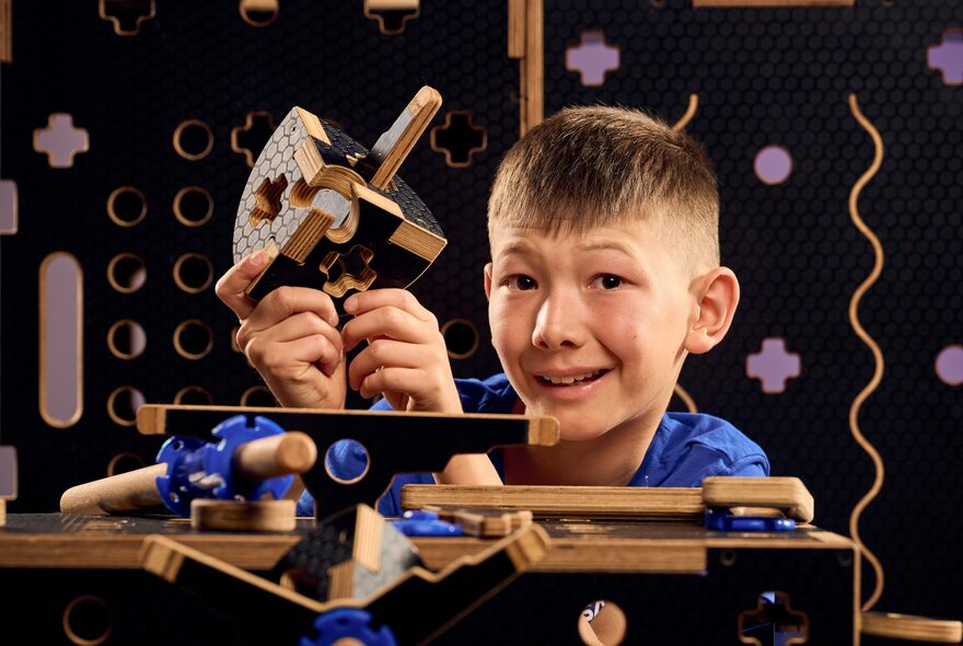 A young boy smiling in front of a timber construction kit that he also holds part of in his hands.