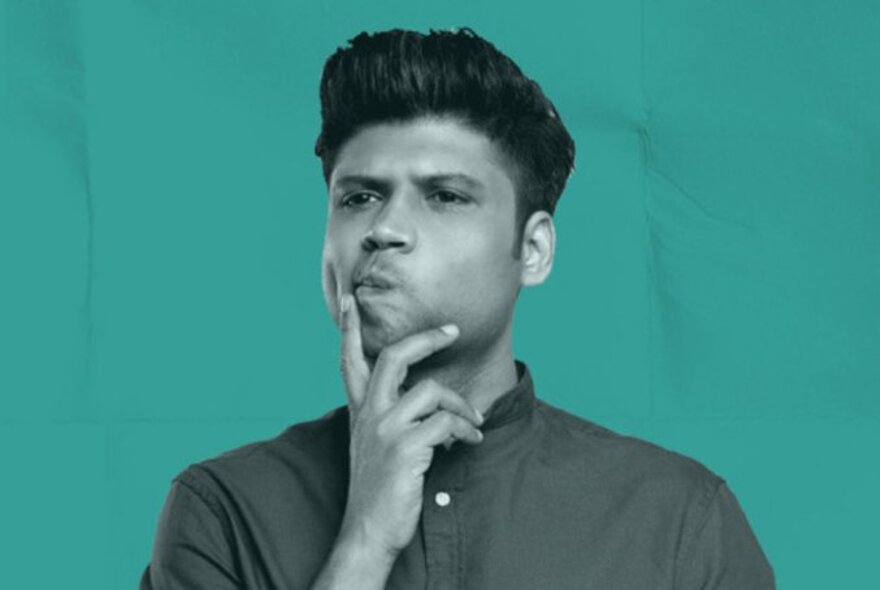 Comedian Rahul Subramanian looking thoughtful with a hand on his chin against a teal background. 