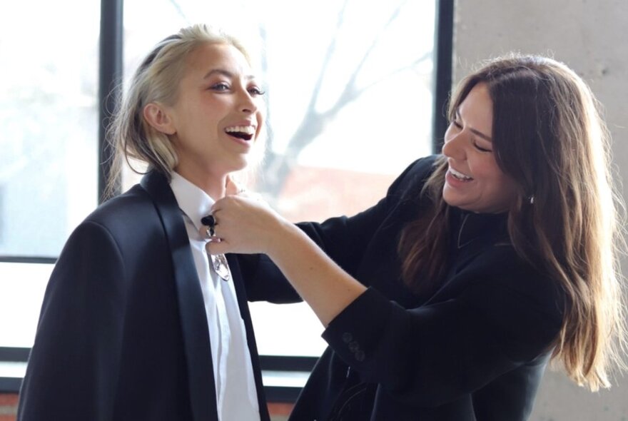 A woman wearing all black straightening a bow tie on a second woman, wearing a black dress jacket and white shirt.