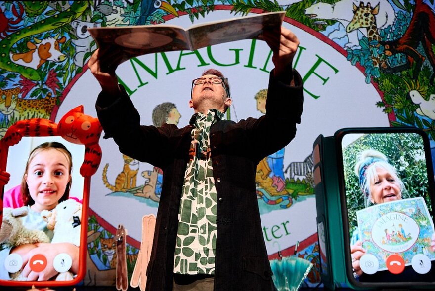 Performer standing onstage next to giant iPhone props, holding a book over their head.