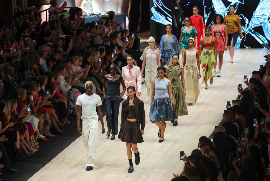 Fashion models walking down a catwalk runway with a seated audience watching, taking photos with their smartphones and applauding.
