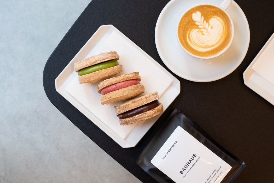 Overhead view of three dacquoise, each with different coloured fillings, on a white plate with a coffee alongside.