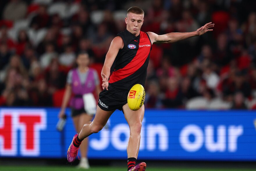 An Essendon AFL football player on the field kicking a yellow football, blurred spectators in the background.