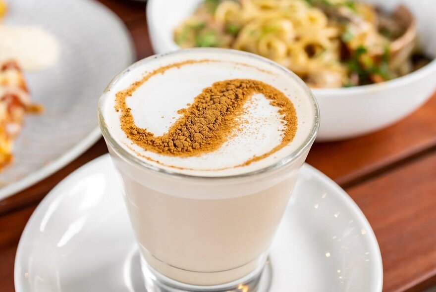 A warm milky drink served in a clear glass mug on a saucer, topped with froth and a decorative dusting of cacao.