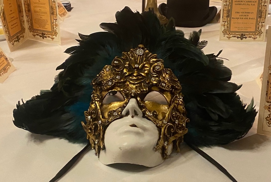 Face sculpture wearing an elaborate feather and gilt mask, lying on a white floor.