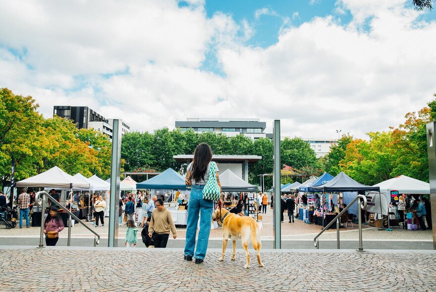 A woman and a golden retriever walking into an outdoor market in a public square.