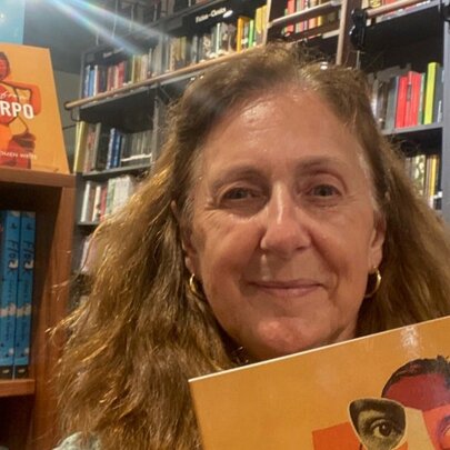 A middle-aged woman holds a book with an orange cover featuring abstract facial artwork, standing inside a bookstore with bookshelves visible in the background.