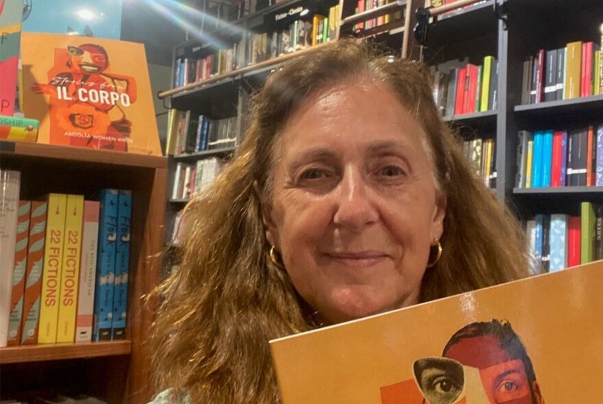 A middle-aged woman holds a book with an orange cover featuring abstract facial artwork, standing inside a bookstore with bookshelves visible in the background.