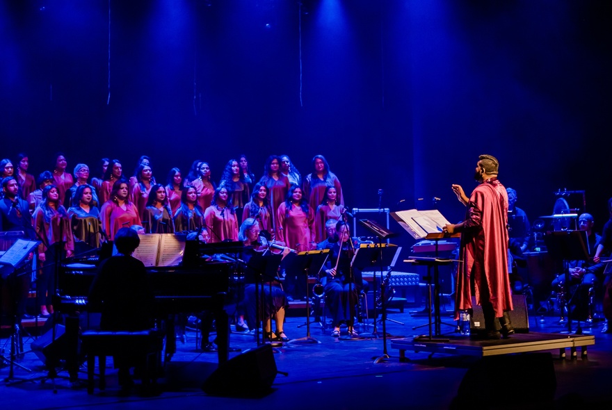 A choir standing on a stage, with a small seated orchestra and a conductor standing in front of them.
