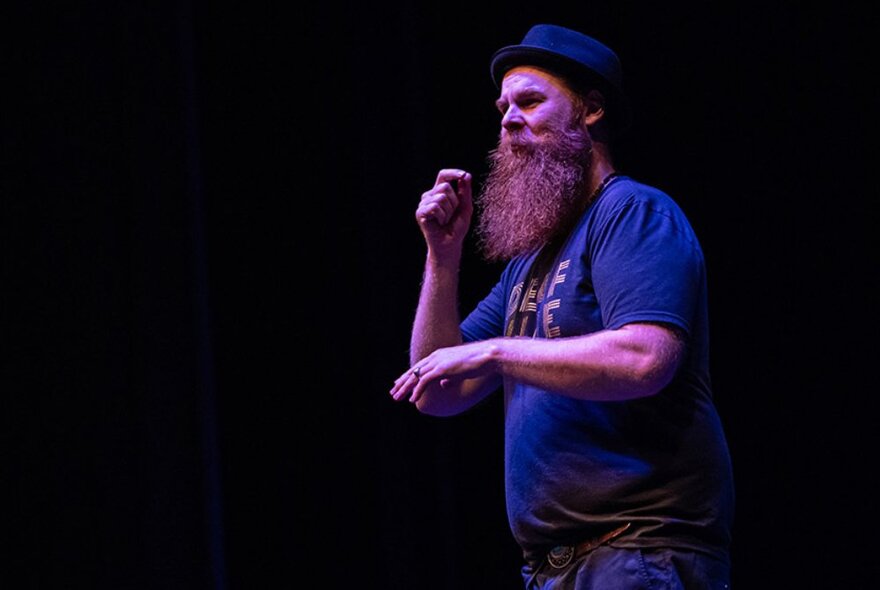 A man with a long beard and wearing a hat, standing on a stage, using his hands to communicate in Auslan.