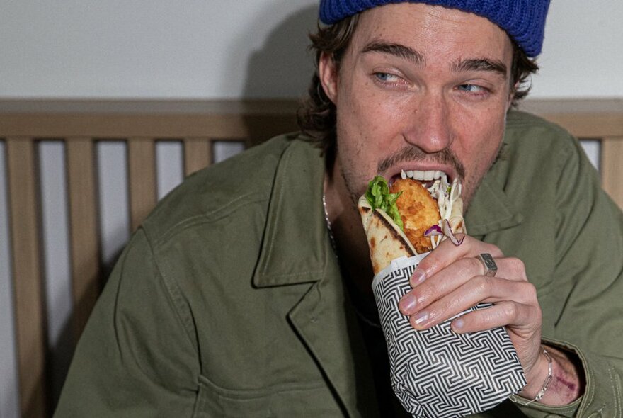 Man with blue beanie biting into souvlaki.