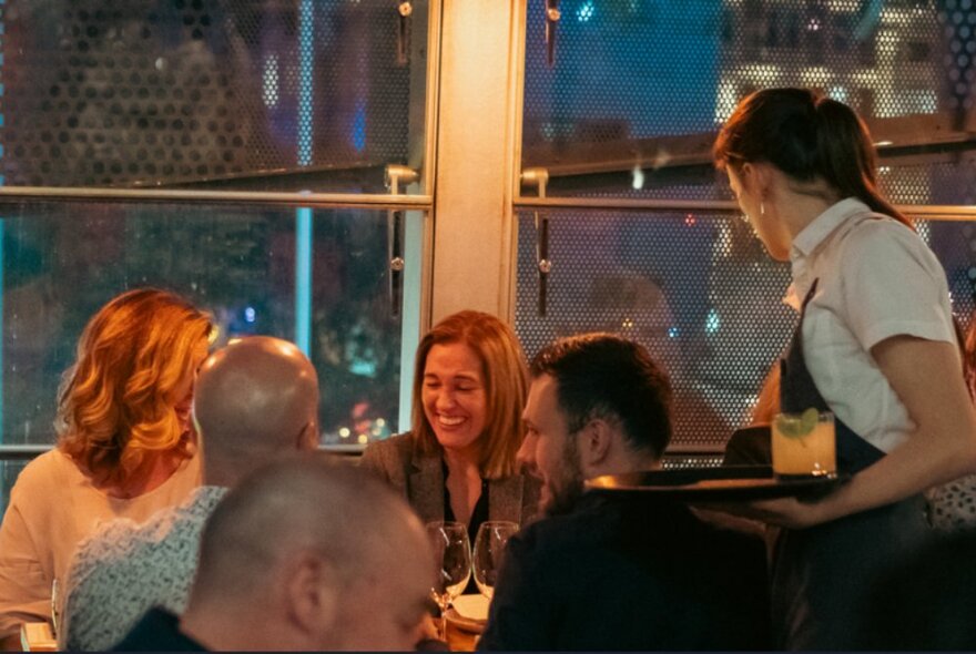 Dining patrons eating and drinking, seated at tables inside a restaurant, with large windows behind them.