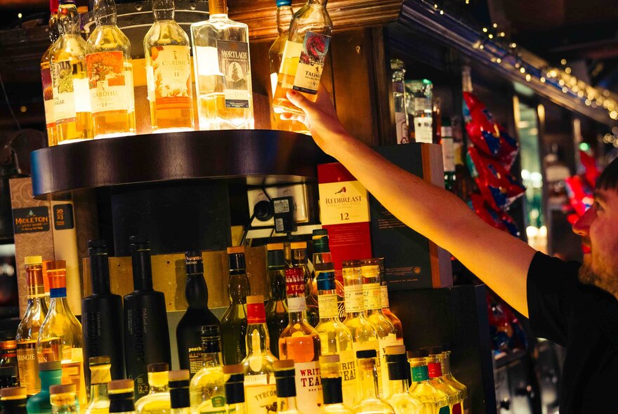 A well-stocked bar with various bottles of spirits and liquors displayed on shelves, and a person reaching up to select a bottle of whiskey from the top shelf.