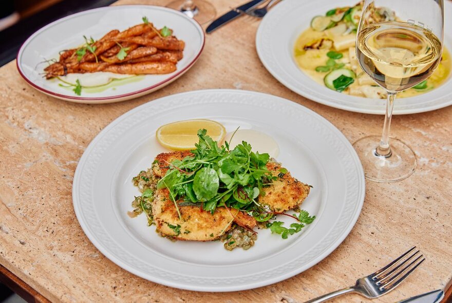 Three white dinner plates with restaurant food arranged on them, and a glass of white wine, on a tabletop.