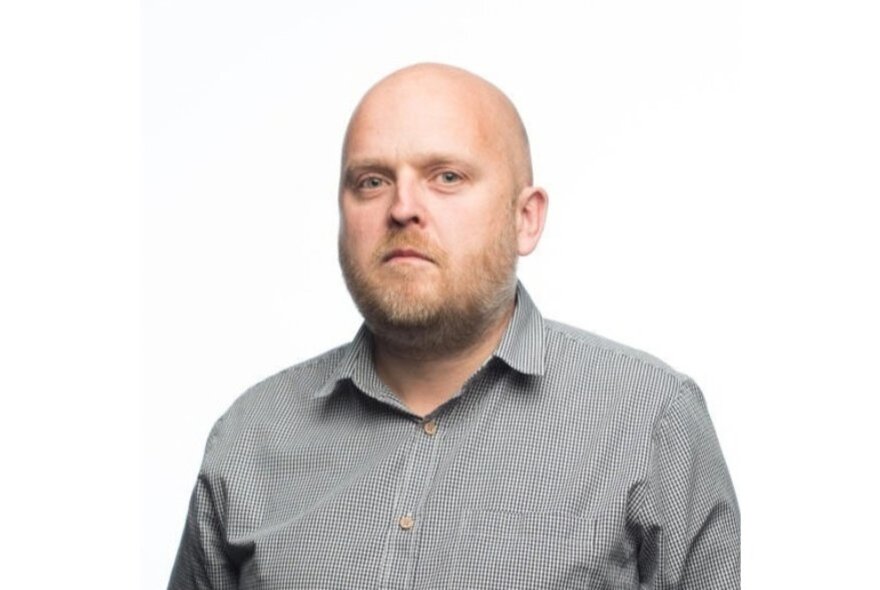 Investigative journalist Ben Schneiders, a bald man with a light beard, wearing a grey long sleeved shirt, posed against a white background.