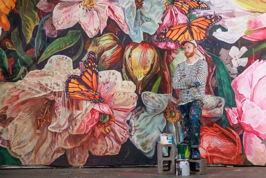 An artist in front a large mural with oversized flowers and butterflies.