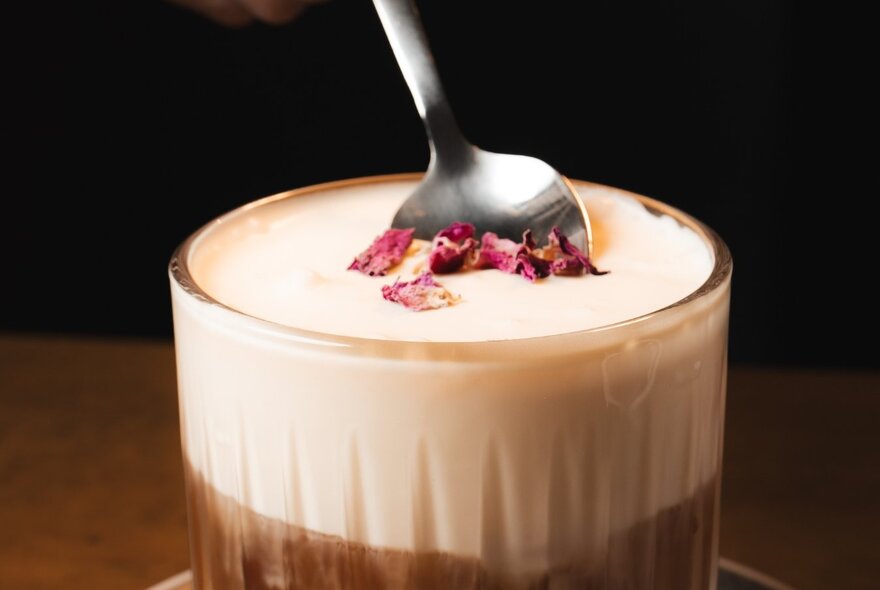 A rose latte coffee, garnished with edible rose petals, with a spoon.