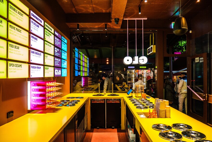 A colourful ice cream shop with a neon-lit menu on the wall and a bright yellow countertop.