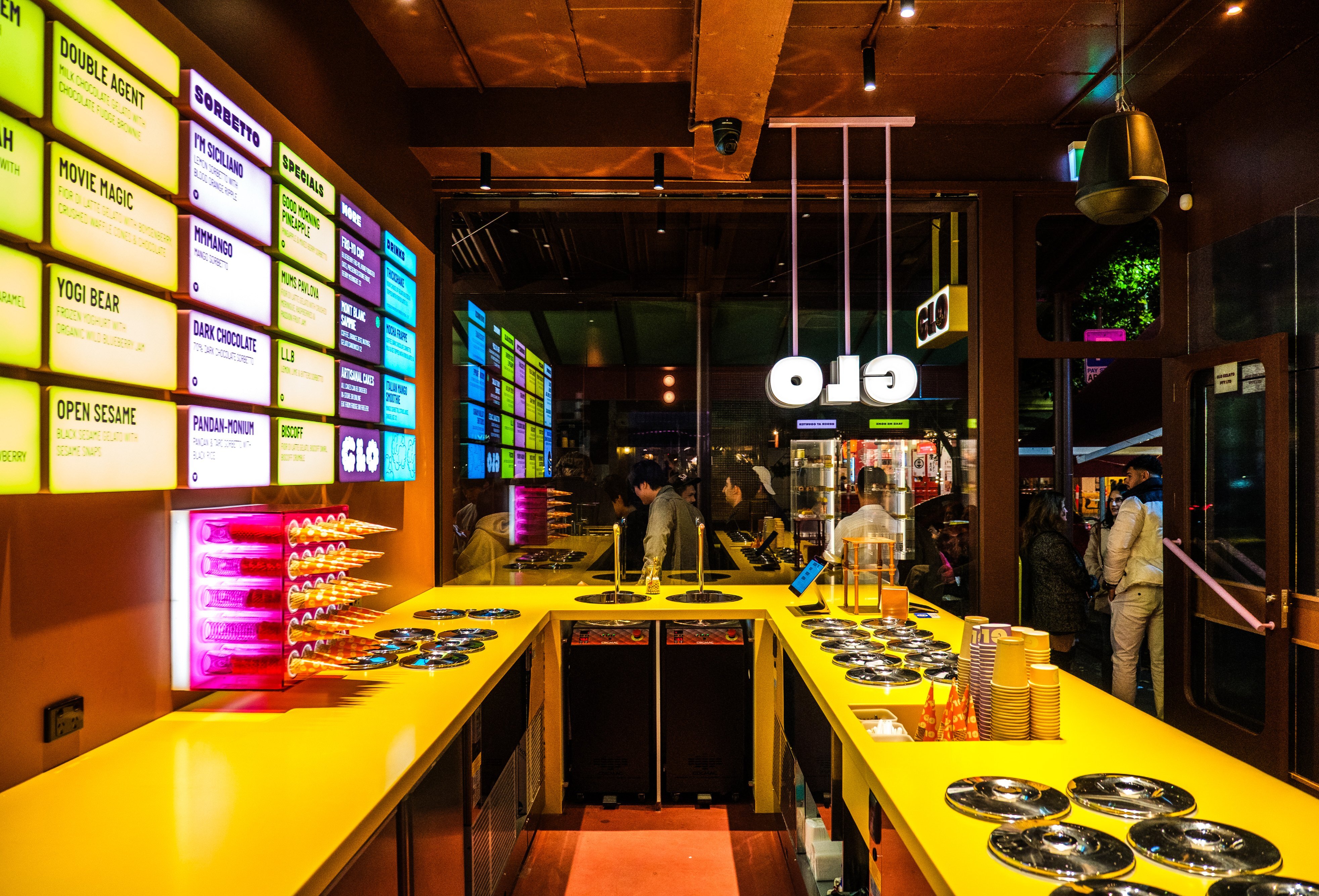 A colourful ice cream shop with a neon-lit menu on the wall and a bright yellow countertop.