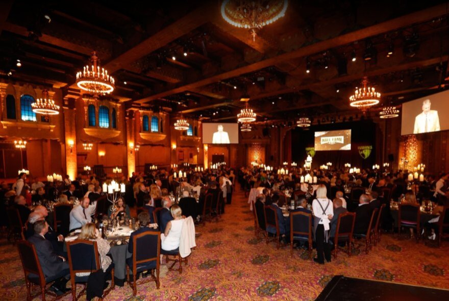 Diners seated at tables in chandelier-lit dining room with several large screens.