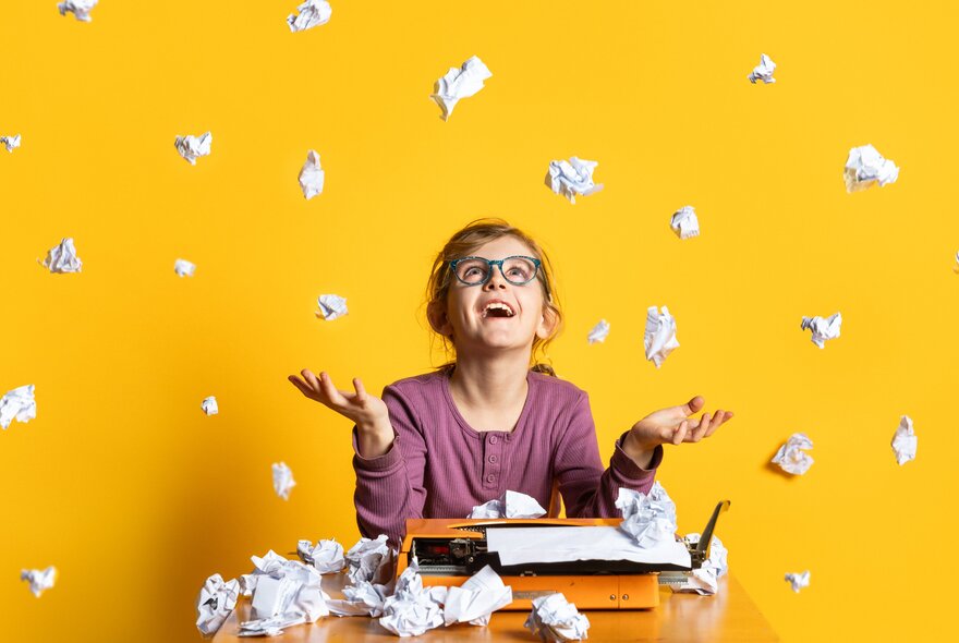A girl throwing bits of paper into the air and smiling in a yellow room, with a typewriter.