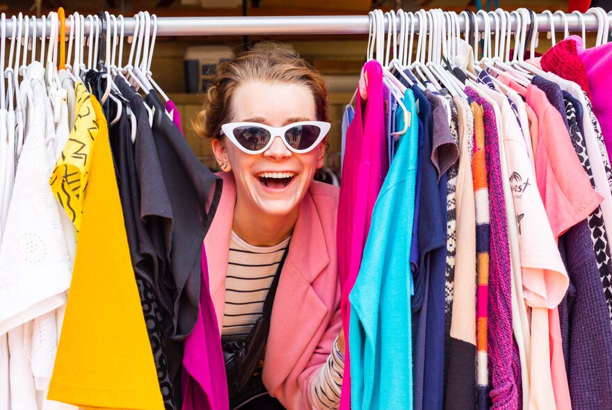 A woman in retro white sunglasses peeking out through clothes hanging on a clothes rail.