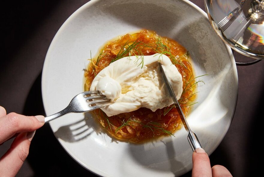 Hands holding cutlery and spreading apart soft cheese on a white plate.