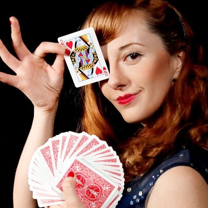 Performer Magical Katrina, holding a pack of  playing cards in one hand, with the Queen of Hearts held up to her face and covering her eye.