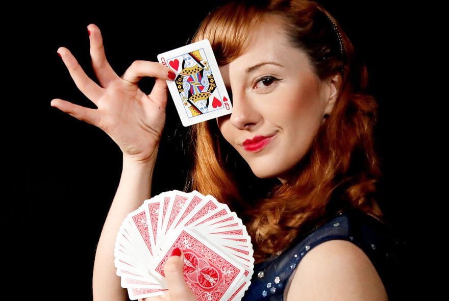 Performer Magical Katrina, holding a pack of  playing cards in one hand, with the Queen of Hearts held up to her face and covering her eye.