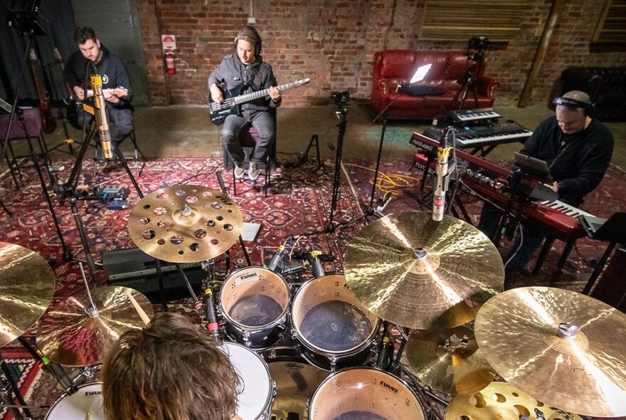 A band playing in a small recording space, looking over the back of the drummer to seated guitarists and a keyboard player.