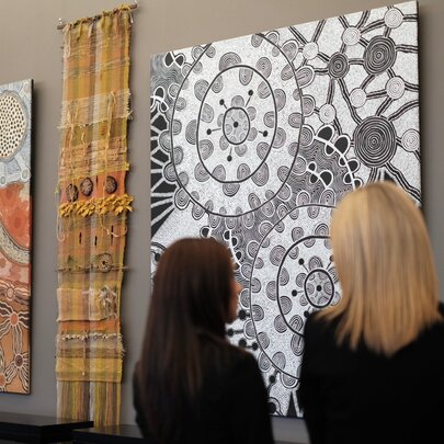Two women viewing large contemporary Indigenous Australian paintings displayed on a wall.