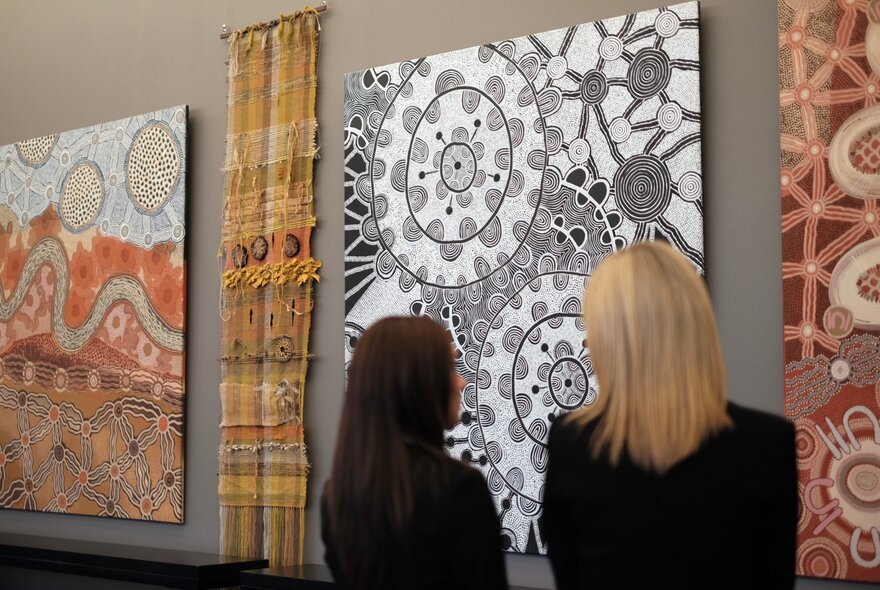 Two women viewing large contemporary Indigenous Australian paintings displayed on a wall.