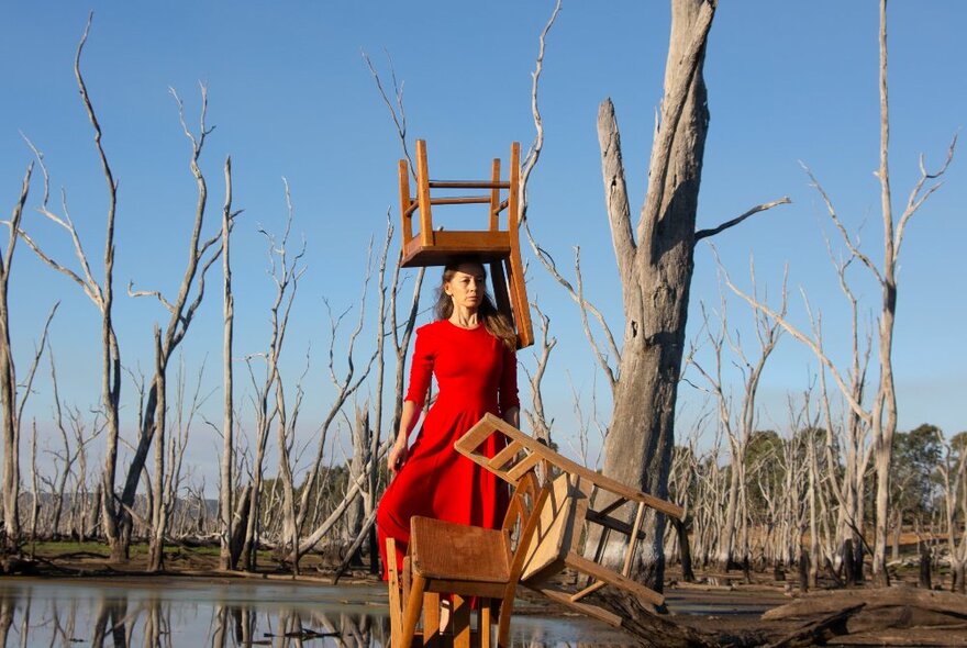 Woman in a red dress, standing in a river bed, dead trees behind her, with a chair balanced on her head, and a few chairs in front of her.