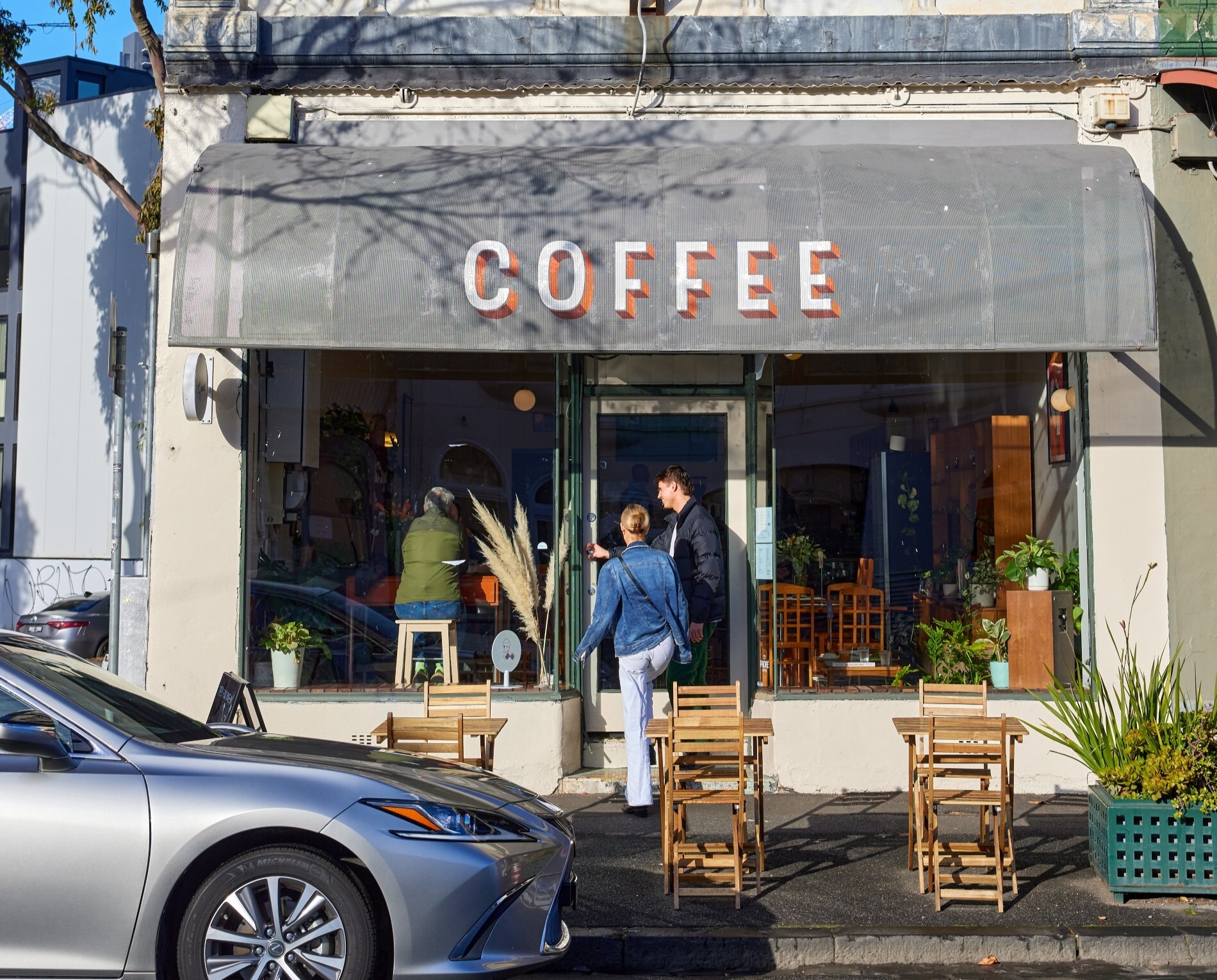 The exterior of a leafy cafe with a black awning and wooden tables and chairs outside