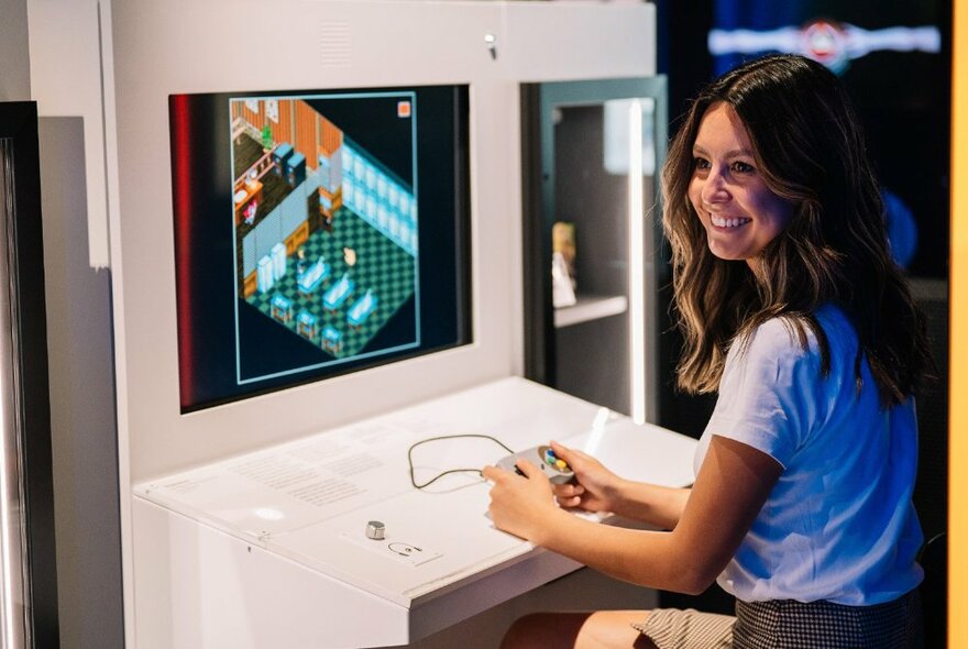 A young woman seated at a white desk playing a game on the screen in front of her, smiling to her left. 