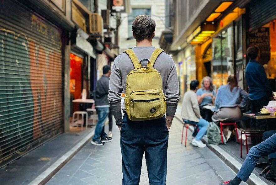 Rear view of a person wearing a green canvas small backpack, standing in a city laneway.