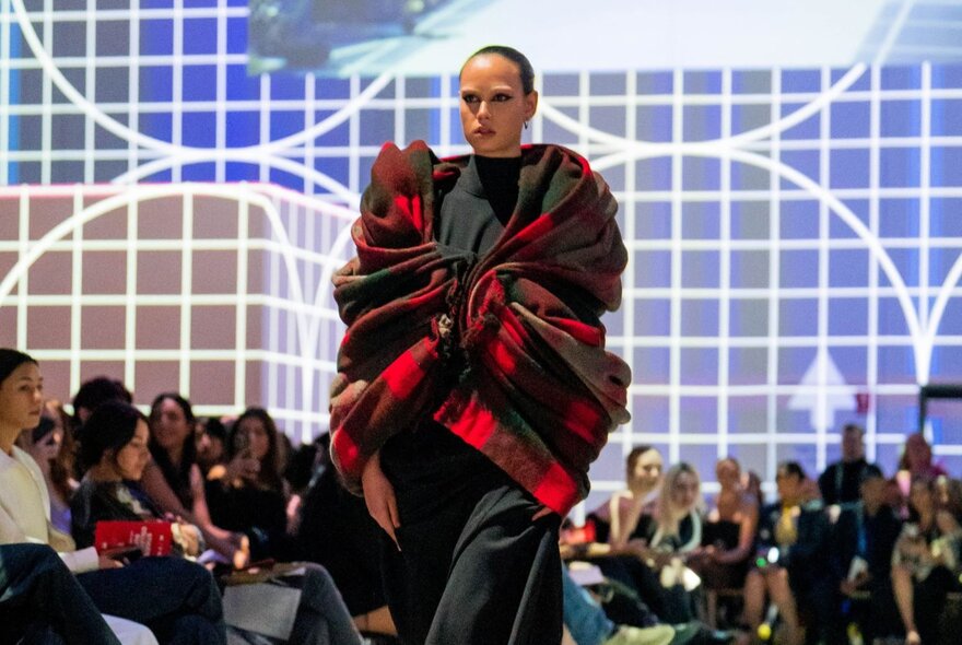 Runway model draped in red tartan stole, with seated onlookers behind her.