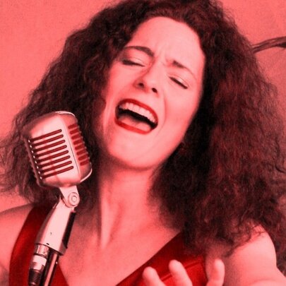 Jazz singer with shoulder-length curly hair, singing into an old-fashioned microphone against red background with plant.
