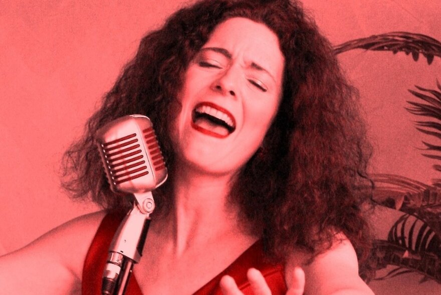 Jazz singer with shoulder-length curly hair, singing into an old-fashioned microphone against red background with plant.