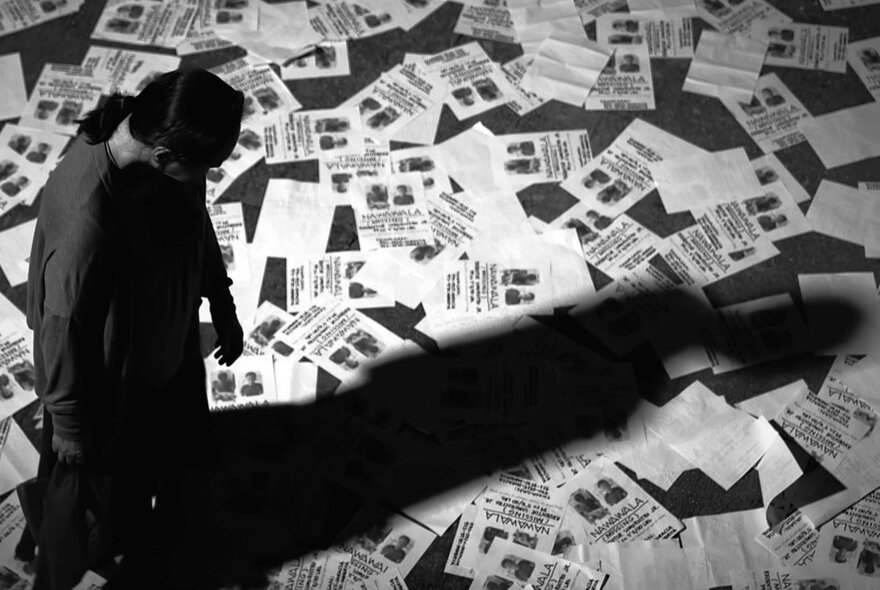Black and white still from the film The Woman Who Left of a person standing on a floor covered in flyers, casting a long, dramatic shadow.