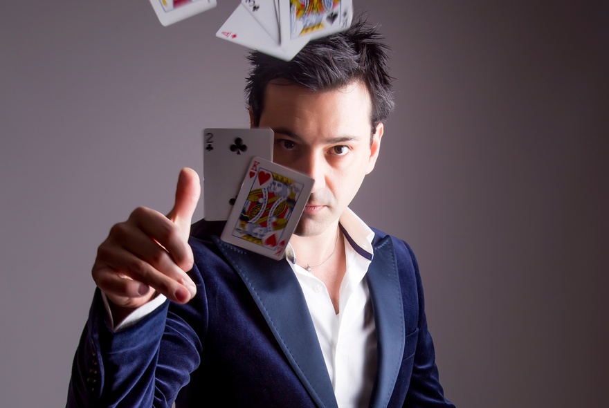 A magician in a blue suit looking directly at the camera while flicking a deck of cards.