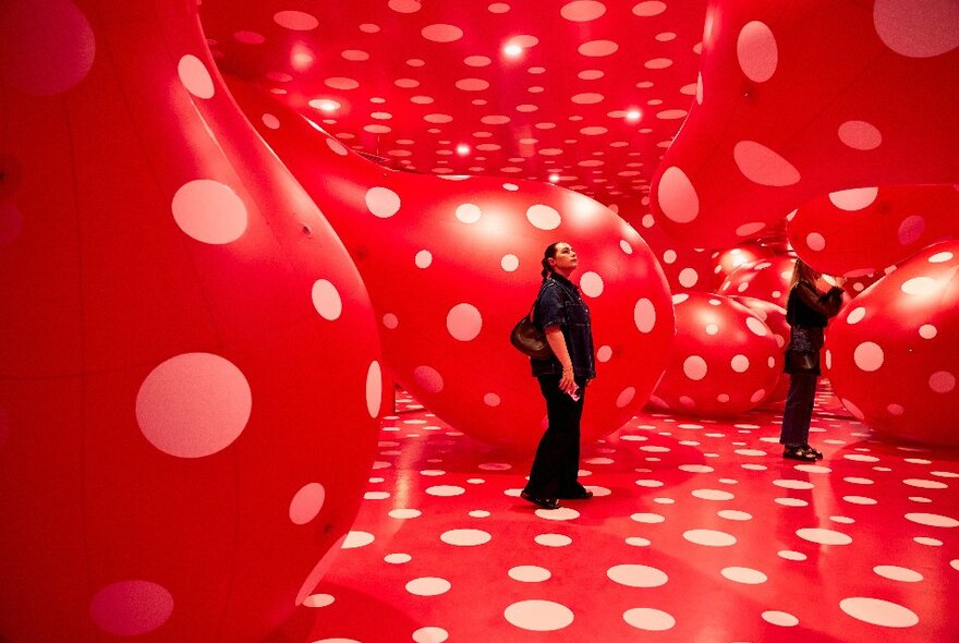 A woman is inside a gallery surrounded by giant red spotty sculptures.