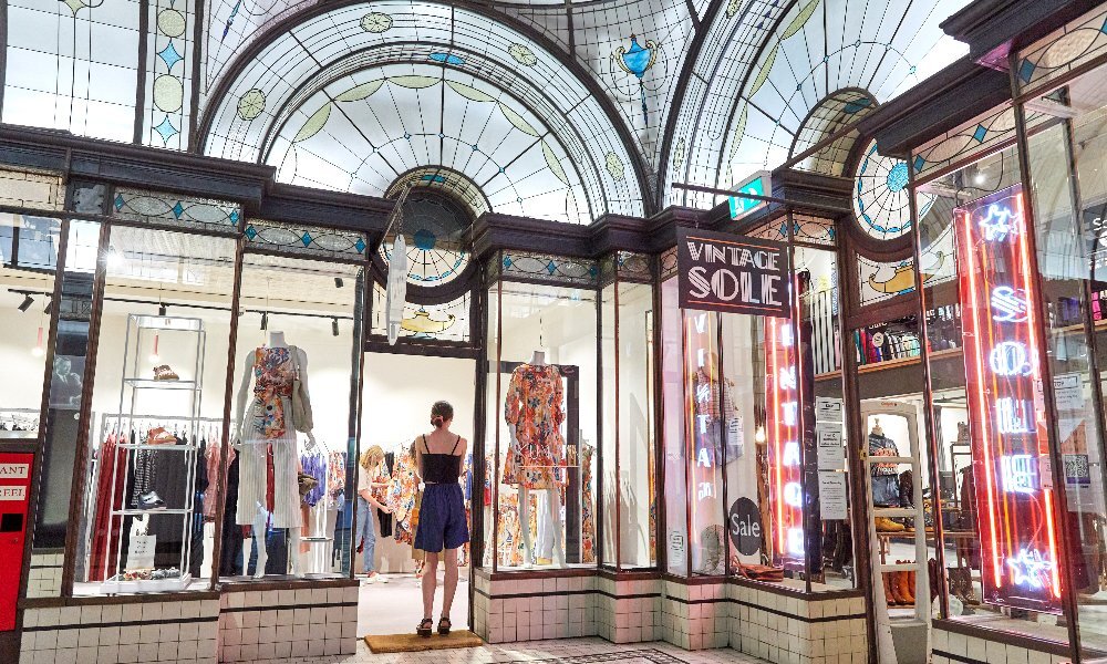 A person standing in the entrance to a clothing boutique inside an Art Deco style shopping arcade.