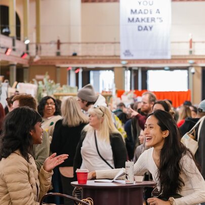 Women interacting over a drink at an indoor market with people browsing in background. 