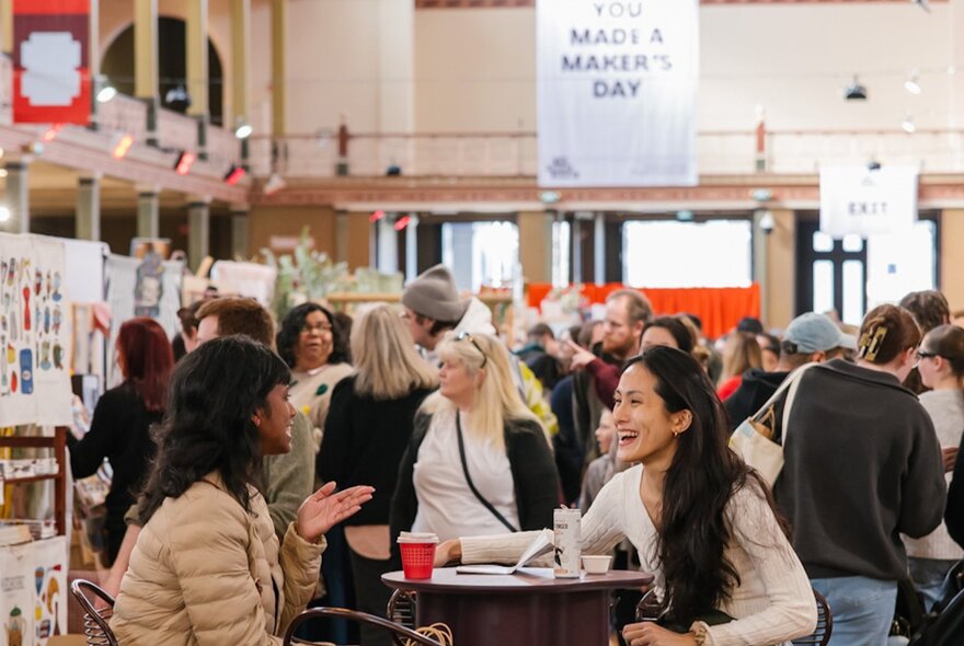 Women interacting over a drink at an indoor market with people browsing in background. 