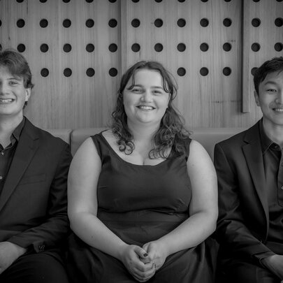 Three young musicians seated together in a row against a wood panelled wall; black and white.