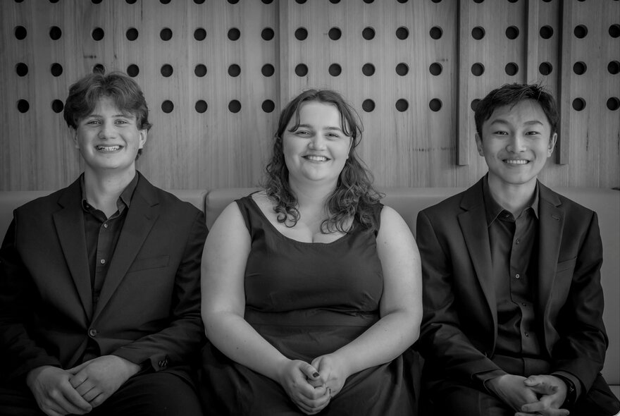 Three young musicians seated together in a row against a wood panelled wall; black and white.