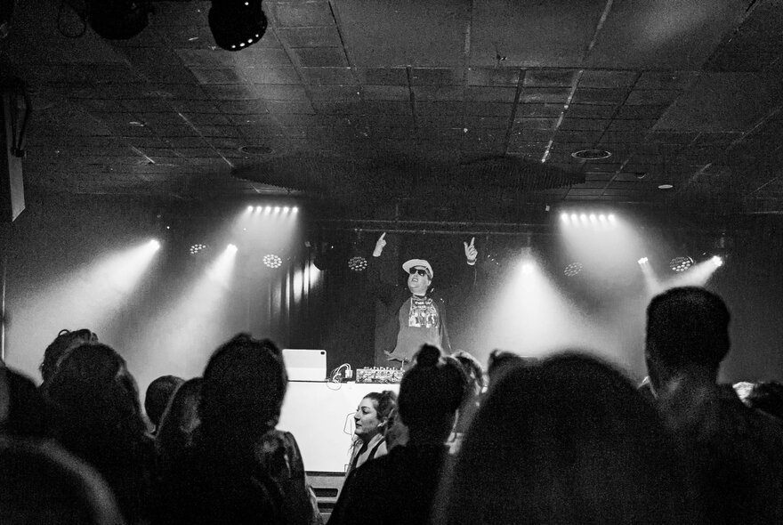 A black and white image of a DJ perfoming a set in a nightclub in front of patrons.